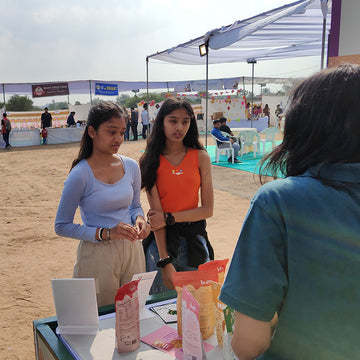 Two women standing at a table with products, with a tent and people in the background.
