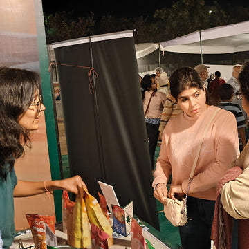 Two women interacting at an outdoor event with a tent and people in the background.
