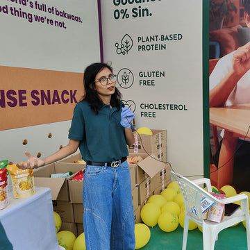 Woman in a green shirt and blue jeans standing in front of promotional banners and boxes with snacks.
